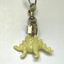 Load image into Gallery viewer, A pale yellow dinosaur keychain charm with a gold-tone metal clasp and a white paper tag, photographed against a plain off-white background.