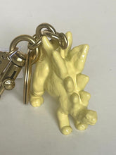 Load image into Gallery viewer, A pale yellow dinosaur keychain featuring a small articulated dinosaur figure attached to gold-tone metal rings and a lobster clasp, shown on a plain light background.
