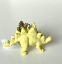 Load image into Gallery viewer, A small pale yellow dinosaur keychain with a metallic ring and attached white paper tag, photographed on a plain light background.