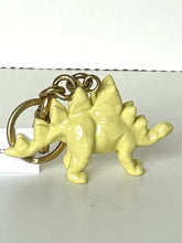 Load image into Gallery viewer, A small pale yellow stegosaurus dinosaur keychain with a gold-tone metal ring and chain, shown on a plain light background.
