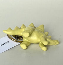 Load image into Gallery viewer, A pale yellow dinosaur keychain charm with a gold-tone metal ring and a white paper tag printed with the brand name COACH.