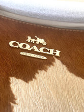 Load image into Gallery viewer, Close-up of a Coach New York logo in gold-tone metal mounted on a brown and beige hair-on-hide surface, with a white fabric trim visible at the top.