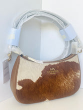 Load image into Gallery viewer, A Coach shoulder bag with a brown and white cowhide pattern, tan leather trim, and a light gray strap wrapped in protective plastic. The Coach logo is visible in gold on the front.