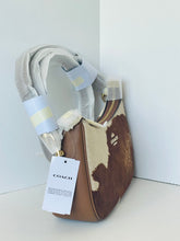 Load image into Gallery viewer, A Coach shoulder bag in brown and cream cowhide with a light gray fabric strap, shown with its hangtag and protective plastic wrapping in a minimalist white corner setting.