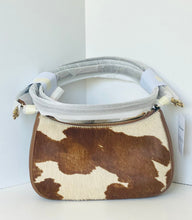 Load image into Gallery viewer, A small shoulder bag with a brown and cream cowhide pattern, featuring a light gray padded shoulder strap and gold-tone hardware, photographed against a white corner wall.