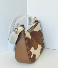 Load image into Gallery viewer, A shoulder handbag with a brown and cream cowhide pattern on the front, smooth brown leather on the side, and a white fabric sleeve wrapping around the handle, set against a plain white background.