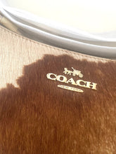 Load image into Gallery viewer, Close-up of a Coach New York handbag made of brown and beige cowhide, featuring a gold-tone metal logo with the horse and carriage emblem.