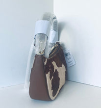 Load image into Gallery viewer, A small shoulder bag with a brown and cream cowhide pattern and solid brown side panels, shown with its white protective plastic wrapping still attached in a white studio corner.