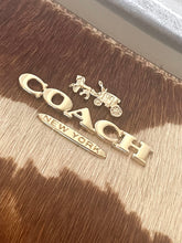 Load image into Gallery viewer, Gold-tone Coach logo charms, including a horse-drawn carriage emblem and a New York City flag-shaped charm, displayed on a brown and beige cowhide surface.