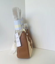 Load image into Gallery viewer, A small brown and beige cow-print shoulder bag with a white hanging tag, standing on a white surface against a white wall. A white towel wrapped in a light blue and white band is leaning behind the bag.