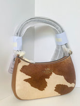 Load image into Gallery viewer, A Coach handbag in brown and cream cowhide, featuring a curved top handle wrapped in protective plastic and a visible gold-tone Coach logo on the front.