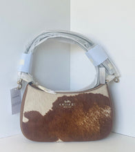 Load image into Gallery viewer, A Coach shoulder bag with a brown and cream cowhide pattern, featuring a silver-tone metal handle and a visible Coach logo on the front, set against a plain white background.