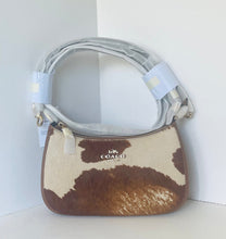 Load image into Gallery viewer, A Coach shoulder bag with a brown and cream cowhide pattern, featuring a light gray adjustable shoulder strap with protective plastic wrapping and a silver-tone logo on the front.