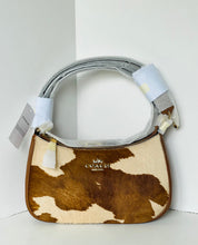 Load image into Gallery viewer, A Coach crescent-shaped shoulder bag with a brown and cream cowhide pattern, featuring a gray faux-leather handle wrapped in protective paper and tags.