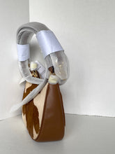 Load image into Gallery viewer, A structured handbag with a brown and cream cowhide pattern body, a thick curved handle wrapped in protective white tape and clear plastic, and a small gold-toned charm, photographed against a plain light gray background.
