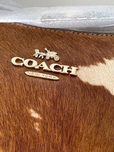 Load image into Gallery viewer, Close-up of a Coach New York metallic logo plaque on a brown and white cowhide surface, with a silver textured material visible at the top.
