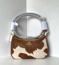 Load image into Gallery viewer, A Coach shoulder bag in brown and cream cowhide with a silver-tone handle, shown in a white corner studio setting. The bag has protective plastic wrapping on the handle and a visible Coach logo on the front.
