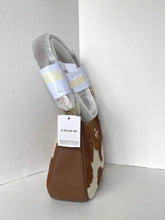 Load image into Gallery viewer, A Coach shoulder bag with a brown and white cowhide pattern and smooth brown leather trim, wrapped in protective plastic and held with white tape. A white COACH hangtag is attached, set against a plain white corner background.