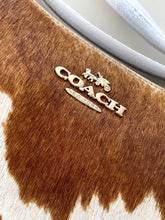 Load image into Gallery viewer, Close-up of a Coach handbag in brown and white cowhide with a gold-tone metal logo plaque on the front.