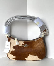 Load image into Gallery viewer, A Coach crescent-shaped shoulder bag in brown and cream cowhide, wrapped in protective gray protective foam and tape on the handle, with a white brand tag hanging from the zipper.
