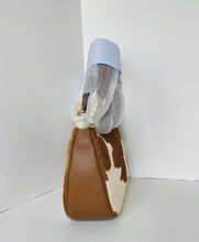 Load image into Gallery viewer, A shoulder bag with a brown and white cowhide pattern and smooth brown leather trim, shown standing upright in a white corner with a clear plastic protective sleeve around the handle.