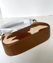Load image into Gallery viewer, A brown faux-leather shoulder bag with a brown and beige cowhide-patterned top flap, resting on a white surface with its strap partially wrapped in protective plastic.