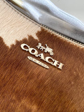 Load image into Gallery viewer, Close-up of a Coach New York handbag featuring a brown and beige cowhide pattern, a silver-tone metallic zipper, and a gold-tone metal logo plaque with the iconic horse and carriage emblem.