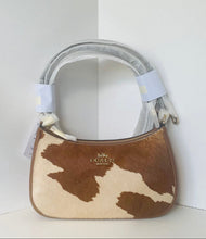 Load image into Gallery viewer, A Coach handbag in brown and cream cowhide, featuring a curved top handle wrapped in protective plastic and white tags attached.