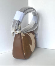 Load image into Gallery viewer, A structured shoulder bag with a brown and cream cowhide patterned body, a solid brown leather front panel, and a wide gray fabric handle, shown with protective white paper wrapping on a white corner shelf.
