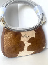Load image into Gallery viewer, A Coach shoulder bag made of brown and cream cowhide, with a light gray protective plastic wrap still attached to the handle, set against a plain white background.