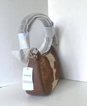 Load image into Gallery viewer, A Coach shoulder bag in brown and beige cowhide with a light gray protective plastic wrap around the handle and a white COACH hangtag, photographed against a plain white background.