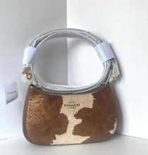 Load image into Gallery viewer, A Coach shoulder bag with a brown and cream cowhide pattern, featuring a light gray handle wrapped in protective plastic and a white COACH tag on the left.