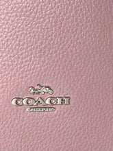 Load image into Gallery viewer, Close-up of a pink pebbled leather surface with a silver-tone Coach logo and horse-and-carriage emblem.