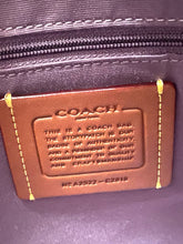 Load image into Gallery viewer, Close-up of a brown leather Coach authenticity patch with gold contrast stitching, sewn onto a dark purple fabric bag near a zipper.