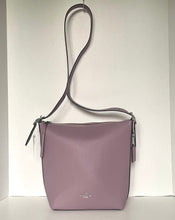 Load image into Gallery viewer, A dusty purple pebbled leather crossbody bag with a matching shoulder strap, hanging against a plain white wall. A small silver-tone Coach logo is visible on the front.