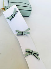 Load image into Gallery viewer, Three light sage green faux leather bow hair clips with black trim, displayed on a white card next to a matching sage green handbag.