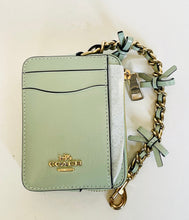 Load image into Gallery viewer, A Coach crossbody wallet in pale sage green leather with gold-tone hardware, featuring a matching green leather chain strap adorned with small bows and a zip-around closure.