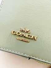 Load image into Gallery viewer, Gold-tone Coach logo charms, including a vertical 'COACH' charm, a horizontal 'CO' charm, and a small 'COCO' charm, displayed on a light sage green textured leather surface.