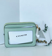 Load image into Gallery viewer, A Coach crossbody bag in light sage green with a clear ID window, gold-tone hardware, and a white branded dust bag beside it on a white background.