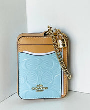 Load image into Gallery viewer, A Coach crossbody card holder in light blue and tan leather with a gold chain and lock, standing on a white surface against a white wall.
