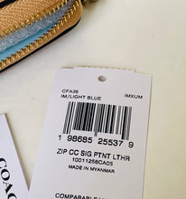 Load image into Gallery viewer, A white COACH zip pouch price tag with a barcode and product code CFA36, shown next to a gold and light blue zipper pouch on a light beige surface.