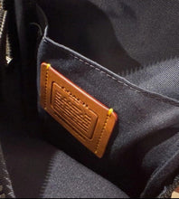 Load image into Gallery viewer, Close-up of the interior of a dark navy or black zippered bag, showing a brown leather brand patch sewn into the lining. A hand is visible holding the bag at the bottom left.