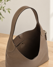Load image into Gallery viewer, A taupe leather shoulder bag with a single top handle, shown from a three-quarter angle on a light wood table with a neutral background.