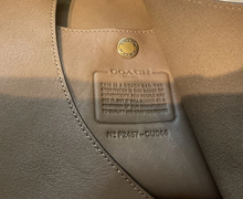 Load image into Gallery viewer, Close-up of a Coach leather interior tag with a gold-tone circular logo and embossed product information on a taupe leather surface.