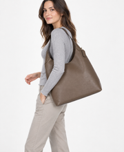 Load image into Gallery viewer, A woman wearing a gray long-sleeve top and beige pants carries a large taupe-brown leather hobo shoulder bag against a plain white studio background.