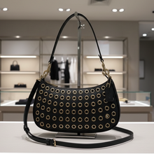 Load image into Gallery viewer, Black handbag with gold studs on a white surface with decorative plants in the background