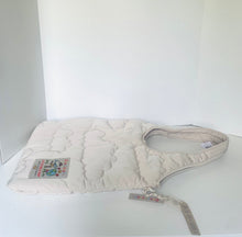 Load image into Gallery viewer, A quilted cream-colored baby carrier or soft pouch with a zippered closure and shoulder strap, resting on a white surface against a white background.