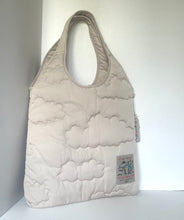 Load image into Gallery viewer, A light beige quilted puffer tote bag with cloud-like stitched patterns, standing on a white surface against a white wall. A small brand patch is visible on the lower right side.