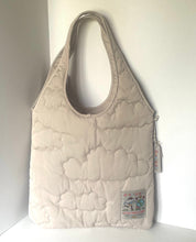 Load image into Gallery viewer, A quilted beige shoulder bag with a cloud-like stitched pattern, standing upright against a plain white background. It features a small decorative patch on the lower right and a zipper closure on the side.