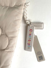 Load image into Gallery viewer, A close-up of a light beige quilted fabric item with a drawstring and attached Coach tags, including a vertical Coachtopia tag and a smaller Coach tag.